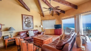 A spacious living room with a large brown leather sofa, a wooden coffee table, and a ceiling fan. There is a large window with a view of the ocean. - Casa Puesta del Sol – Beachfront, Rocky Point (Puerto Peñasco)