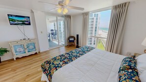 Ein Hotelzimmer mit Bett, Fernseher, Deckenventilator, Blick auf den Strand und einem Balkon mit Stuhl.