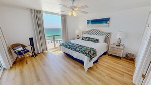 Ein Schlafzimmer mit Bett, Deckenventilator, Nachttisch, Lampe, Korbstuhl und Blick auf den Strand.