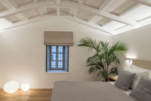 A bedroom with a sloped ceiling, a window with a beige blind, a large potted plant, a bed with gray bedding, and two spherical light fixtures on the floor. - NESEA BOUTIQUE APARTMENTS - THETIS (Idra)
