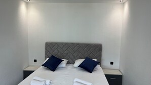 Un lit bien fait, avec du linge de lit blanc et deux oreillers bleu foncé, une table de chevet avec une lampe et un luminaire fixé au mur.