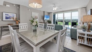 Un espace repas moderne avec une table en verre et des chaises blanches, un ventilateur de plafond et une vue sur l’extérieur grâce à de grandes portes coulissantes.