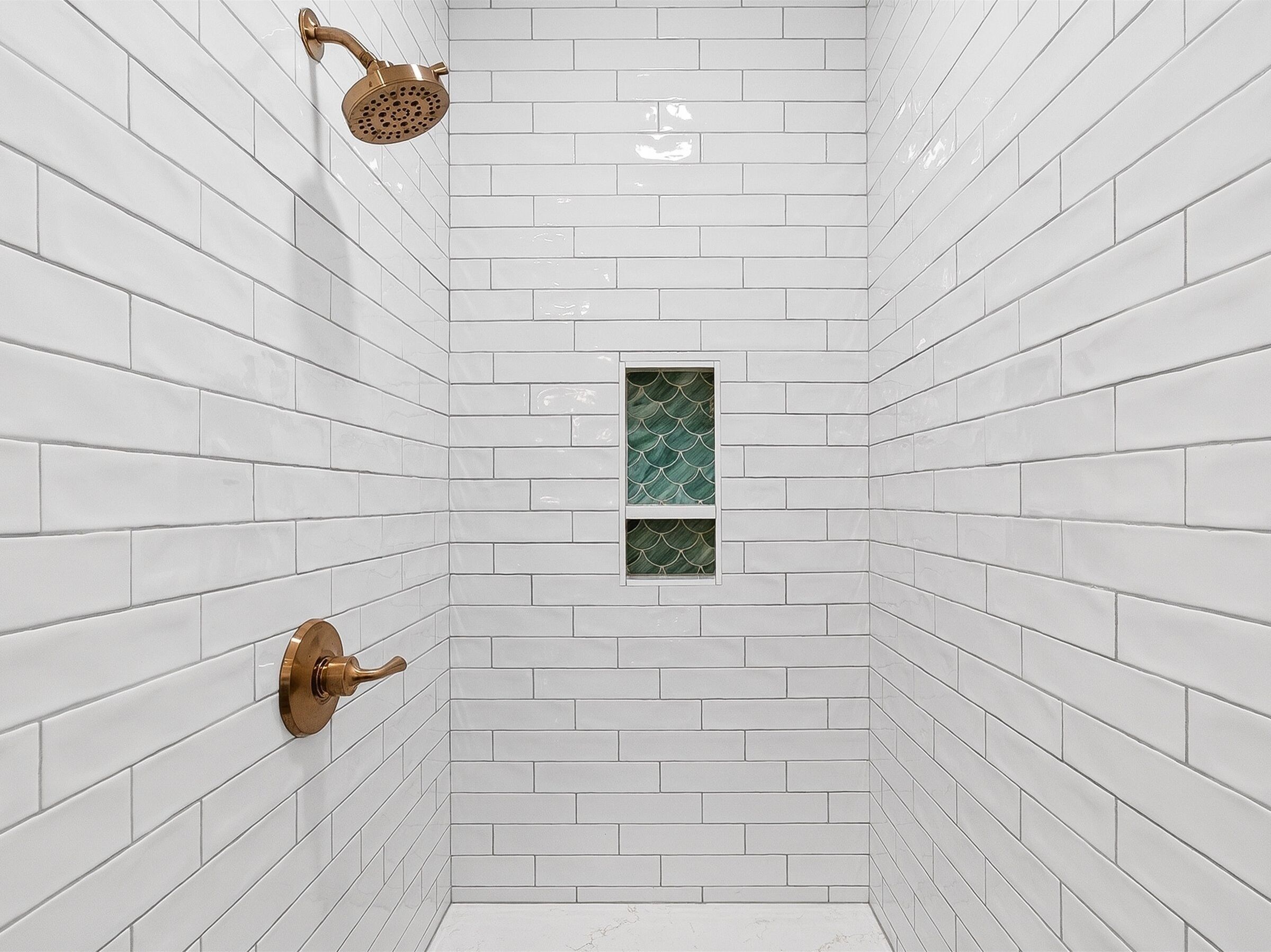 Une salle de bain avec des carreaux de céramique blancs rectangulaires, une petite fenêtre ornée d’un motif vert et une pomme de douche dorée.