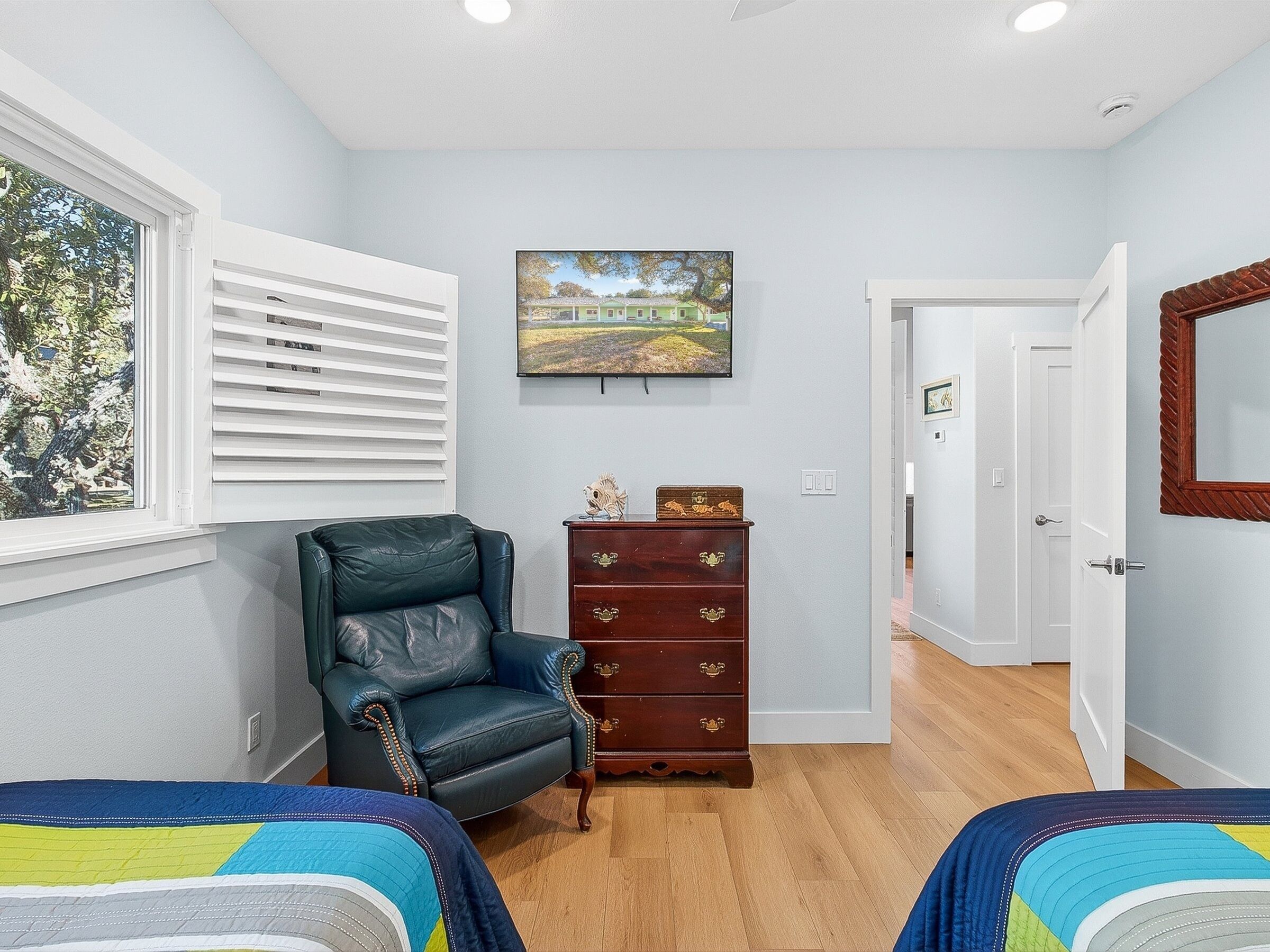 Une chambre avec deux lits, une commode en bois, un fauteuil en cuir, un tableau accroché au mur et une fenêtre avec des stores.