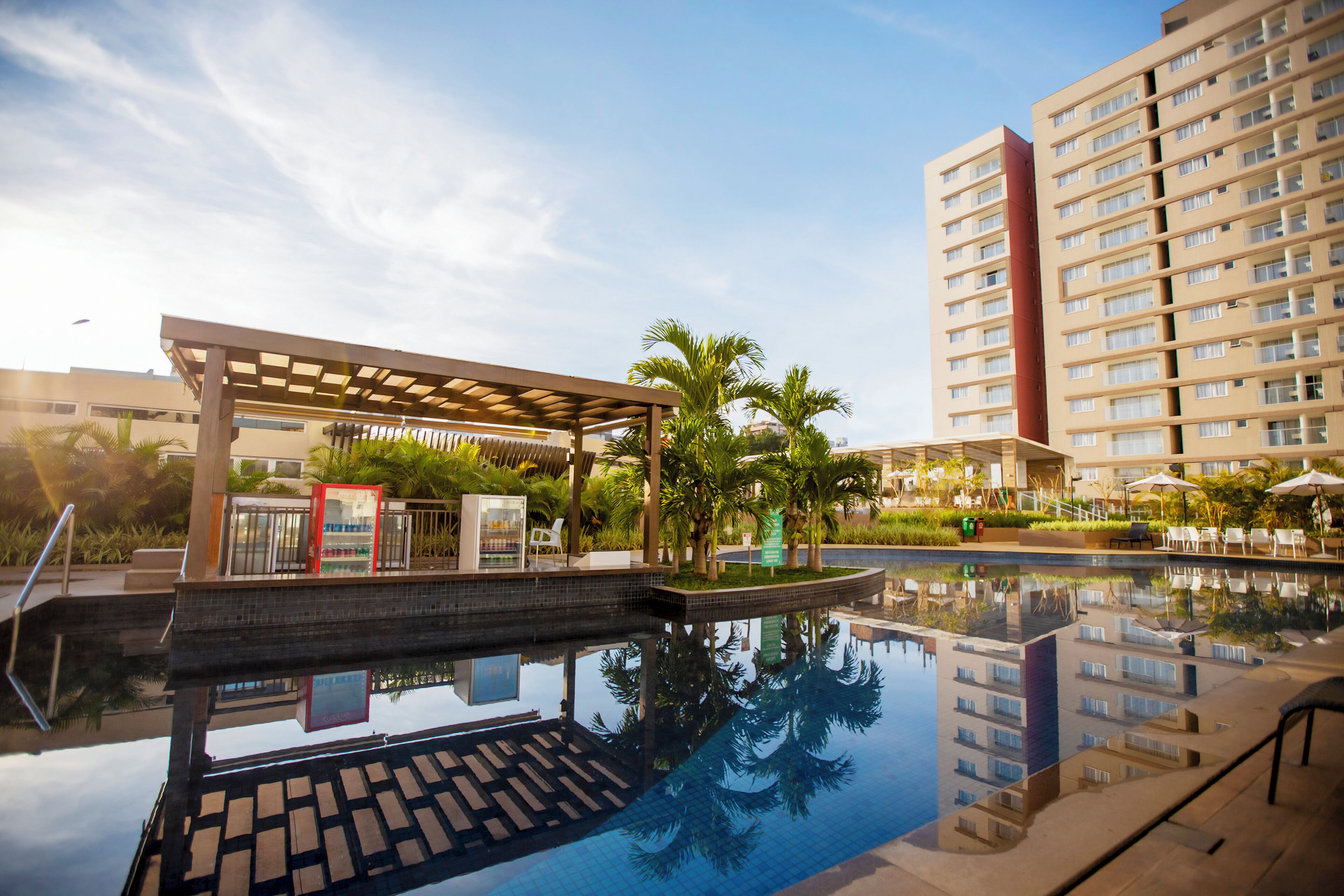 Area piscina di un hotel con una zona salotto coperta, palme e un edificio alto sullo sfondo.