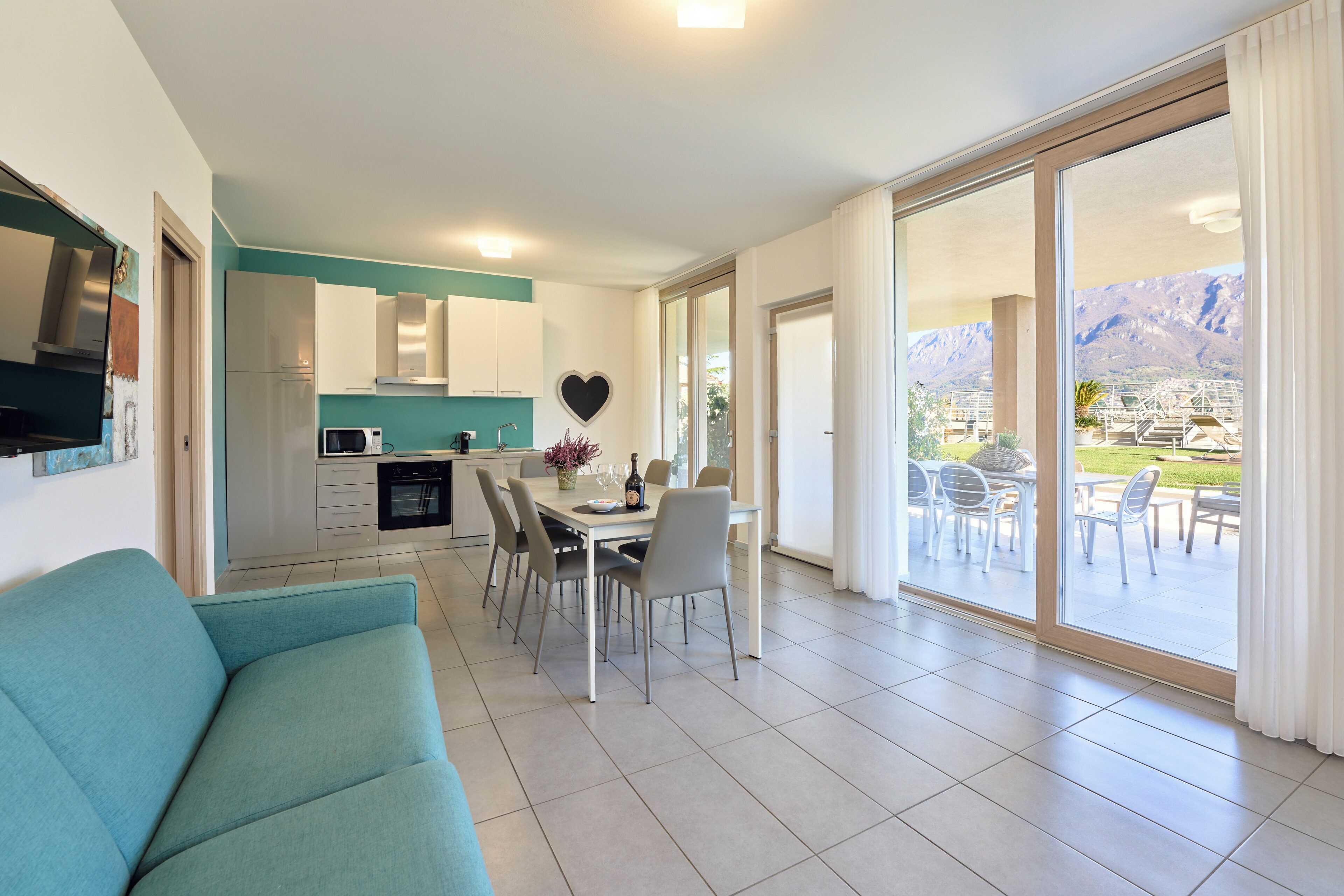 A modern kitchen with white cabinets and a heart-shaped decoration, a dining area with a table and chairs, and a view of mountains through large sliding glass doors.