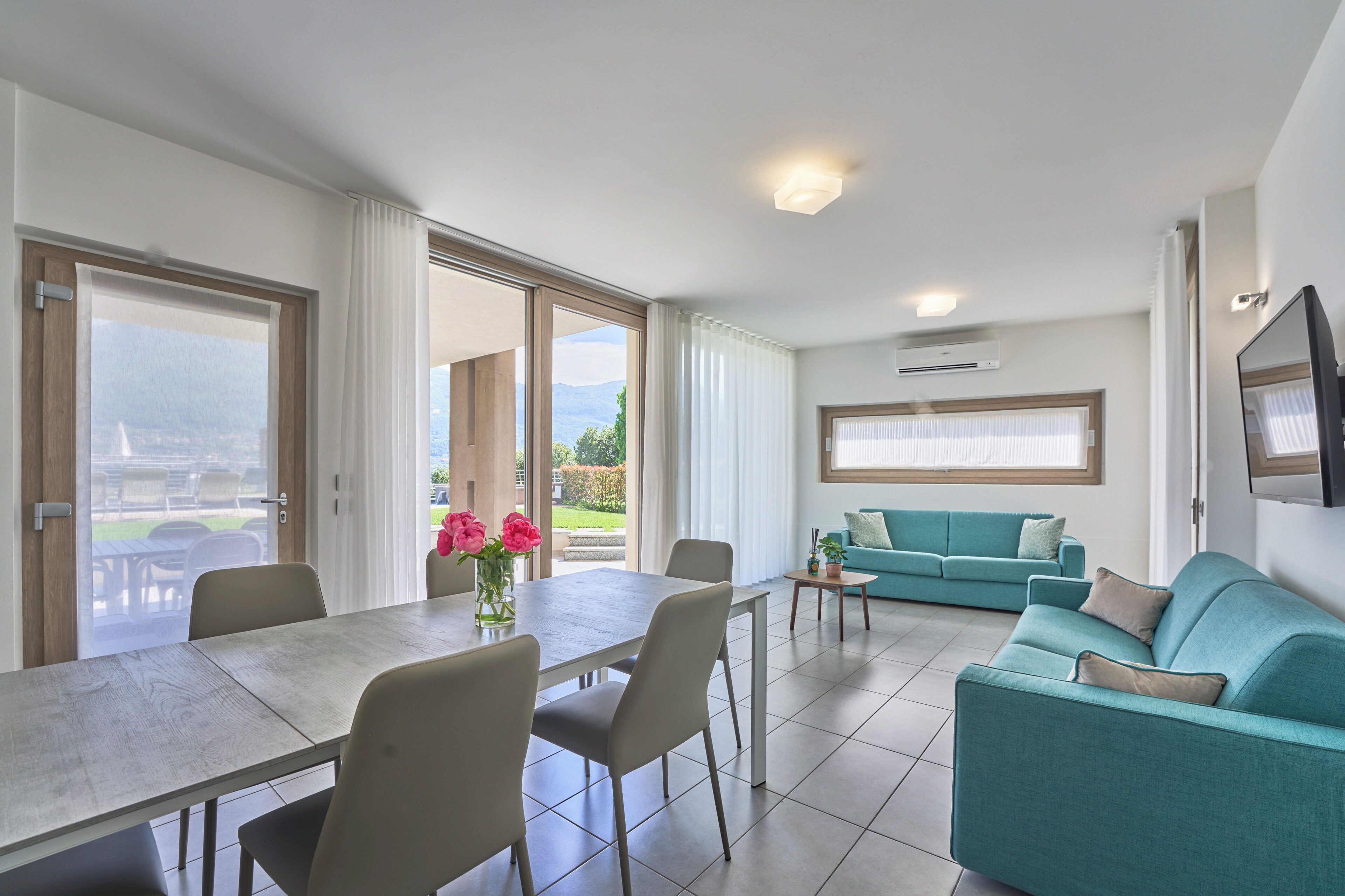 A modern living room with a dining table, teal sofa, and large windows.