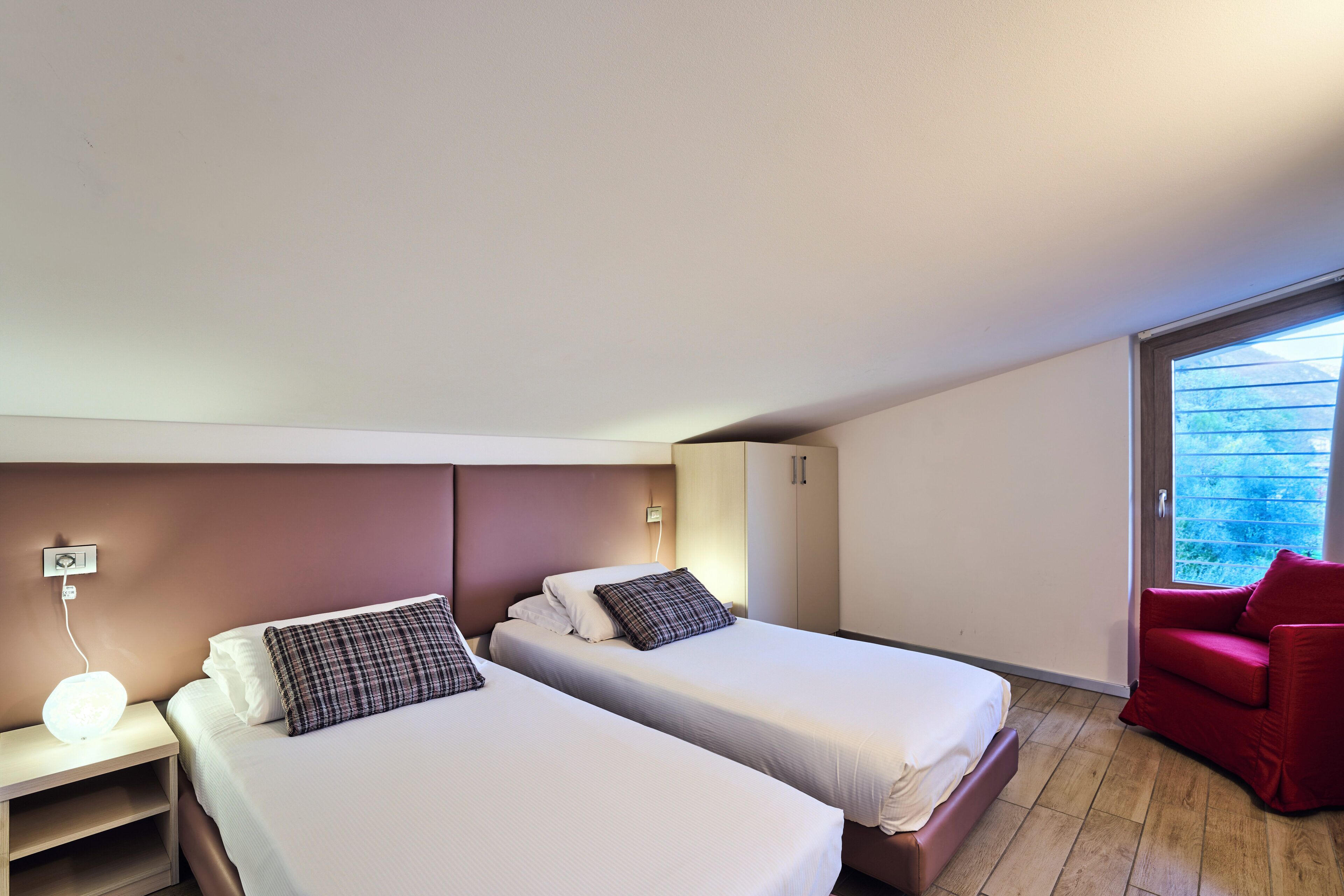 Una habitación de hotel con dos camas, una silla roja, una mesita de noche con lámpara y una ventana con vistas a zonas verdes.