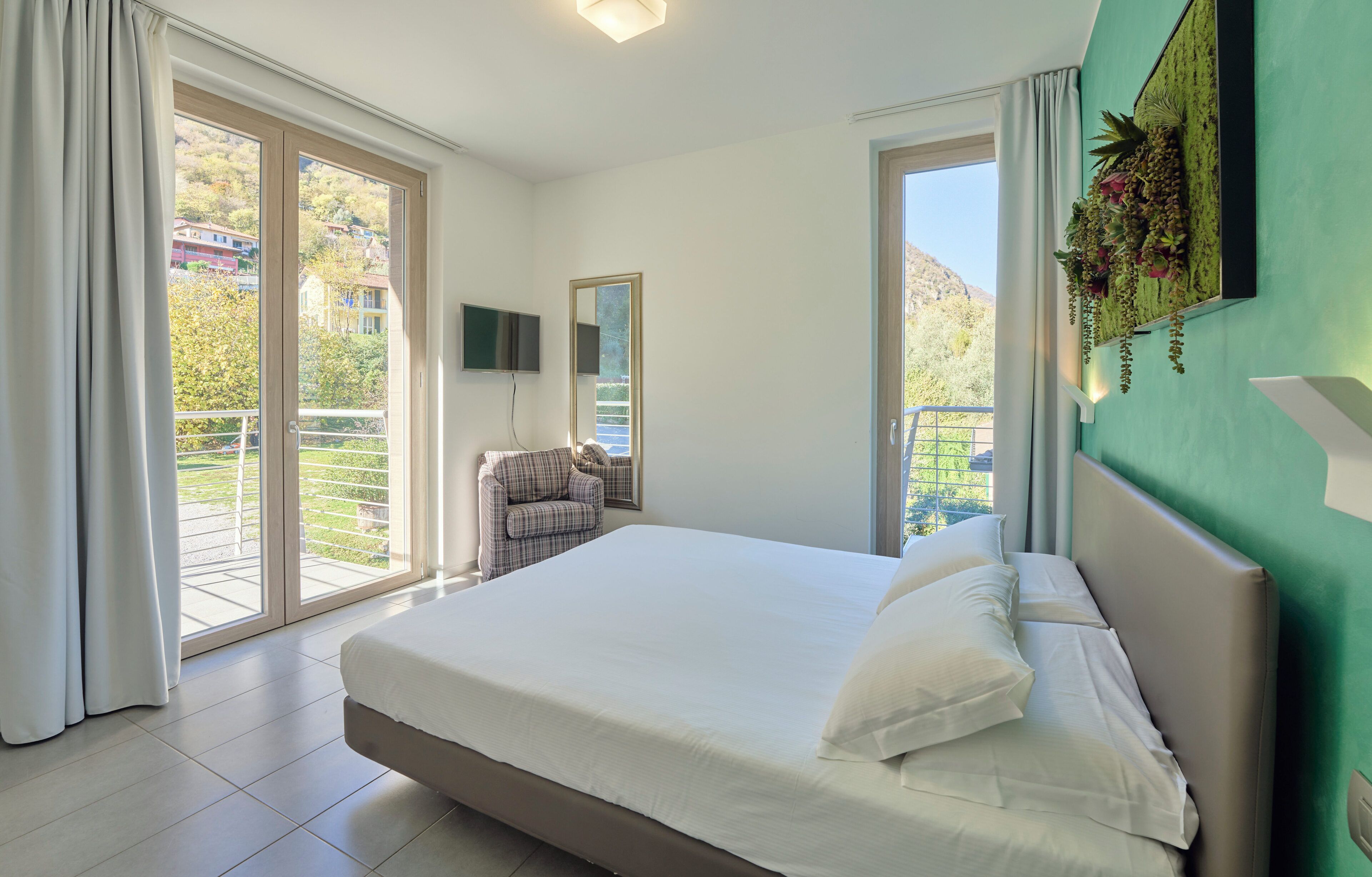 A bedroom with a large bed, a chair, a TV, and a balcony with a view of houses and greenery.