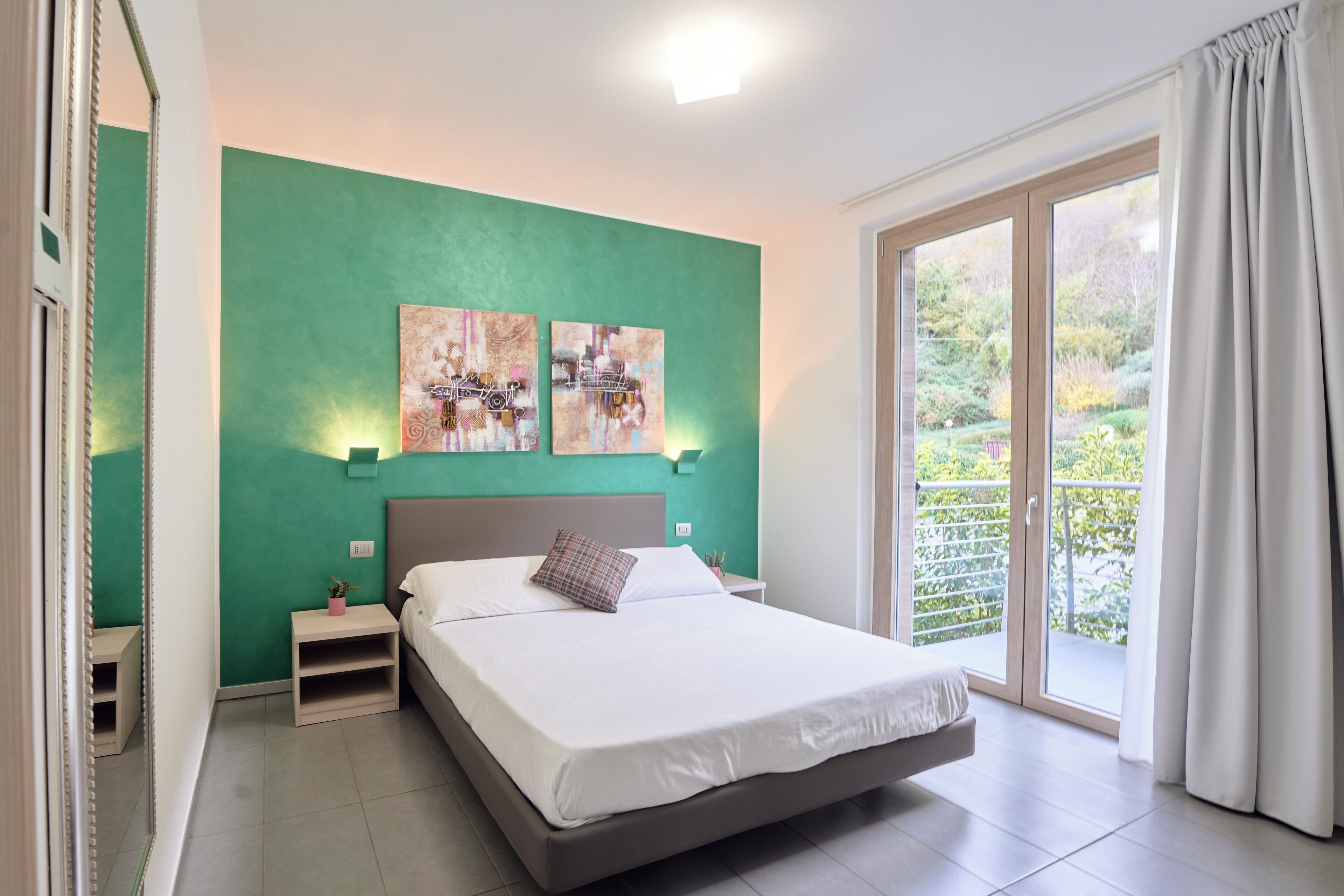 A bedroom with a bed, nightstands, artwork on the wall, and a view through a sliding glass door.