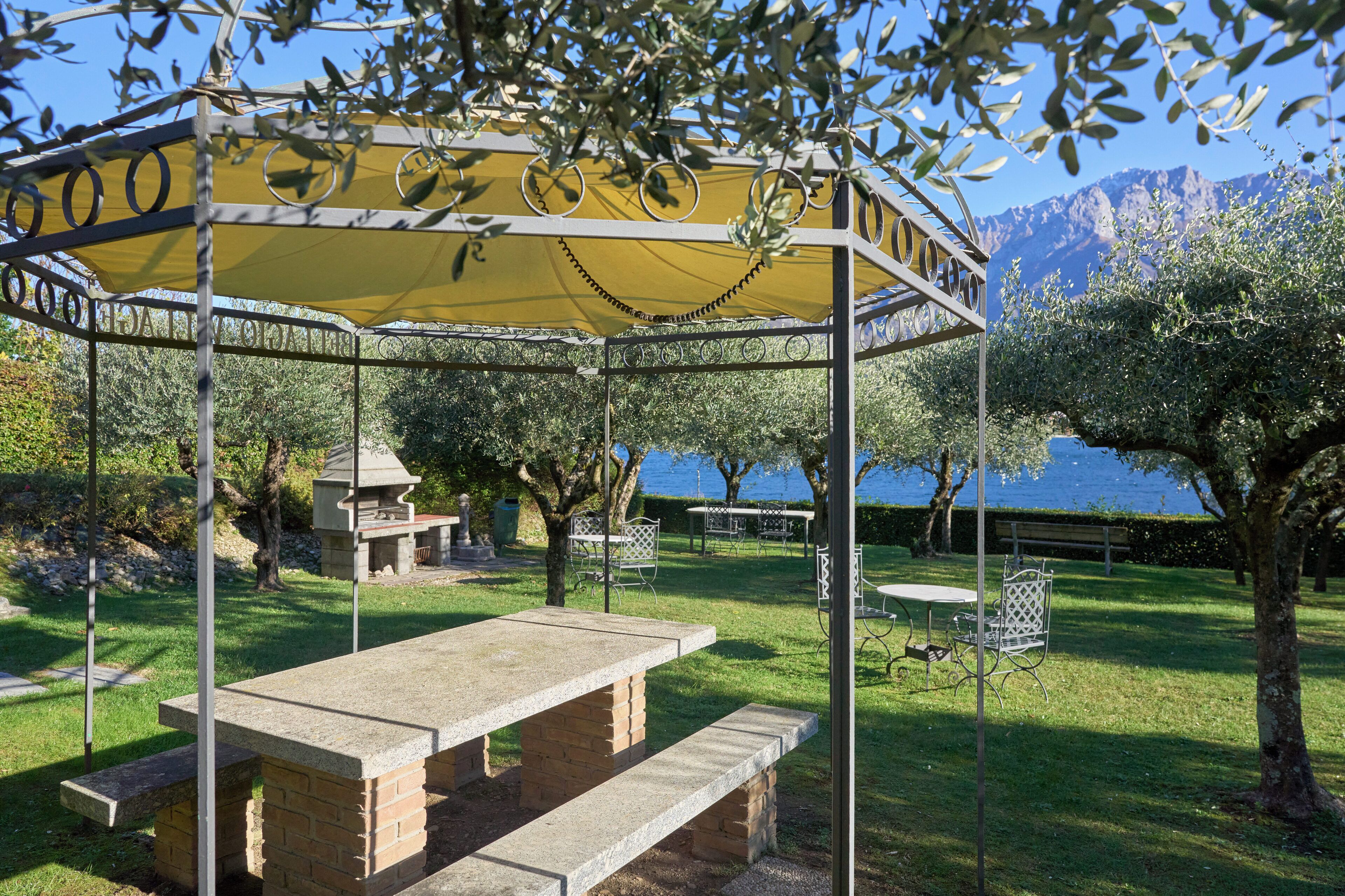 Een tuinpaviljoen met een tafel en banken, omringd door bomen en uitzicht op een meer en bergen.