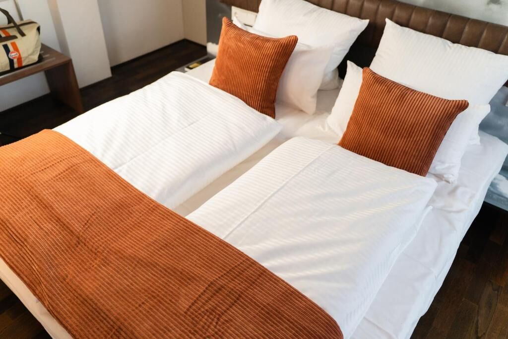 Una cama doble con ropa de cama blanca y naranja, un cabecero de madera y una mesita de noche con un bolso.