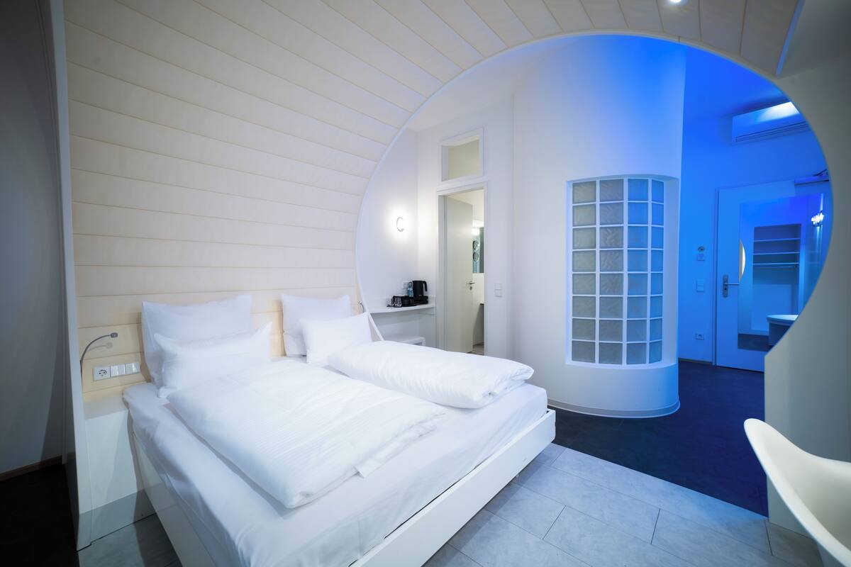 A modern hotel room with a large bed, a circular window, and a view of the hallway.