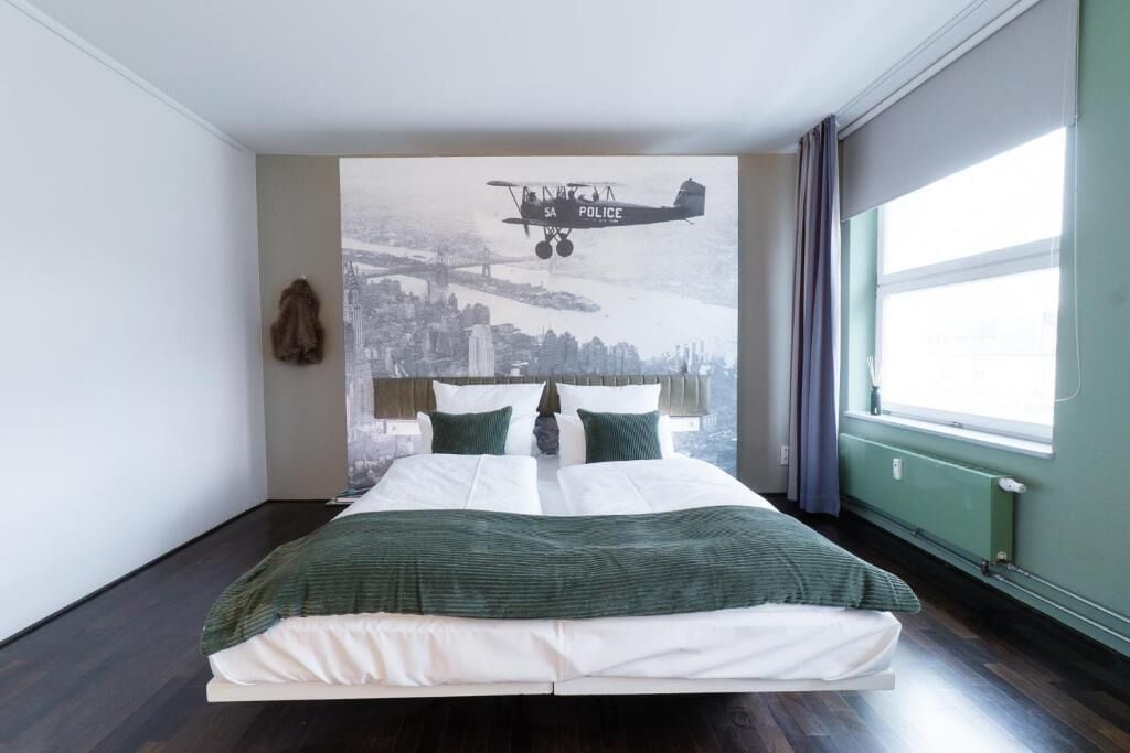 Un dormitorio con una cama grande, un mural de un paisaje urbano en la pared y una ventana con cortinas.