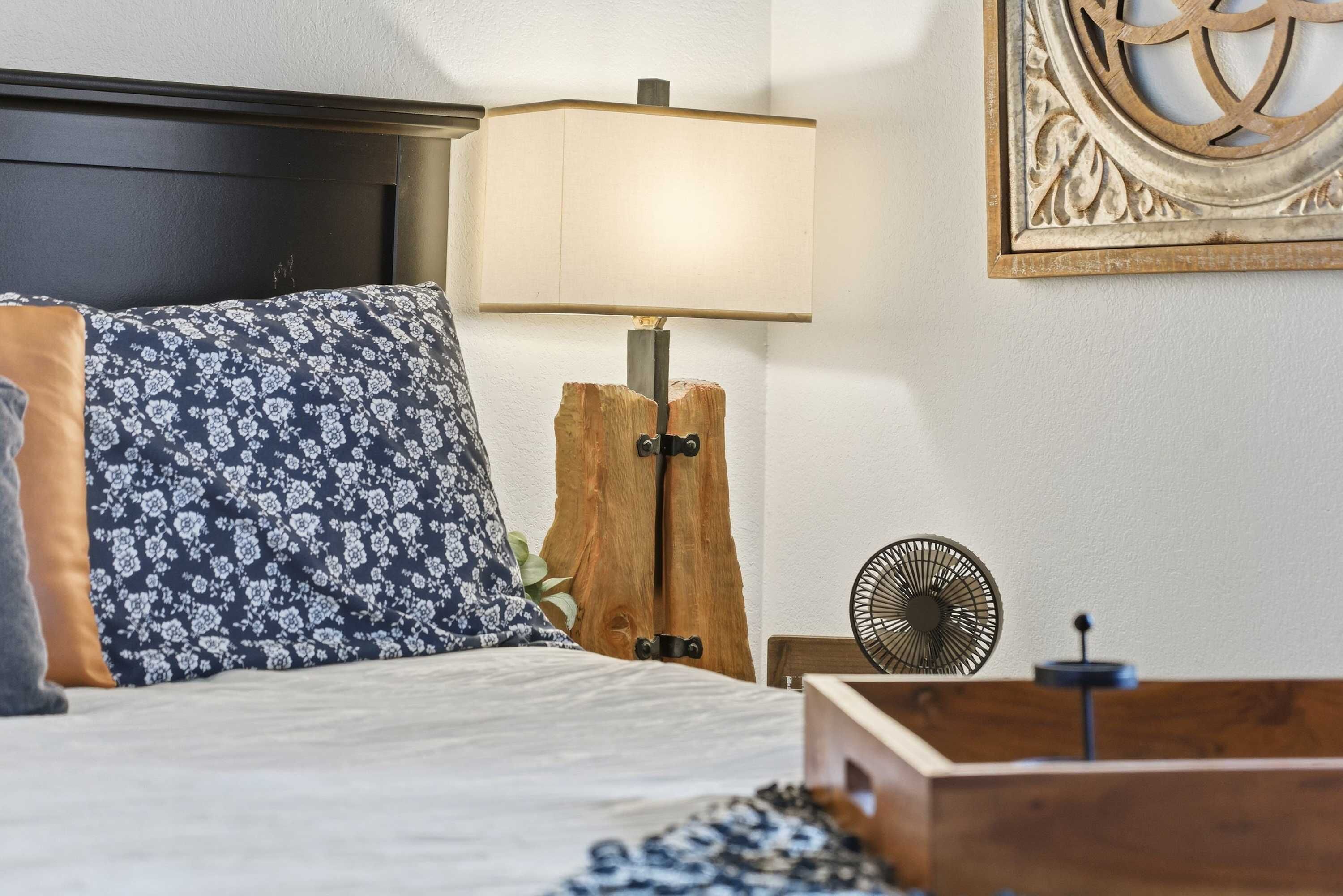 Ein ordentlich bezogenes Bett mit floralen Kissen, ein Nachttisch aus Holz mit einer Lampe, eine Wanddekoration und ein kleiner Ventilator.