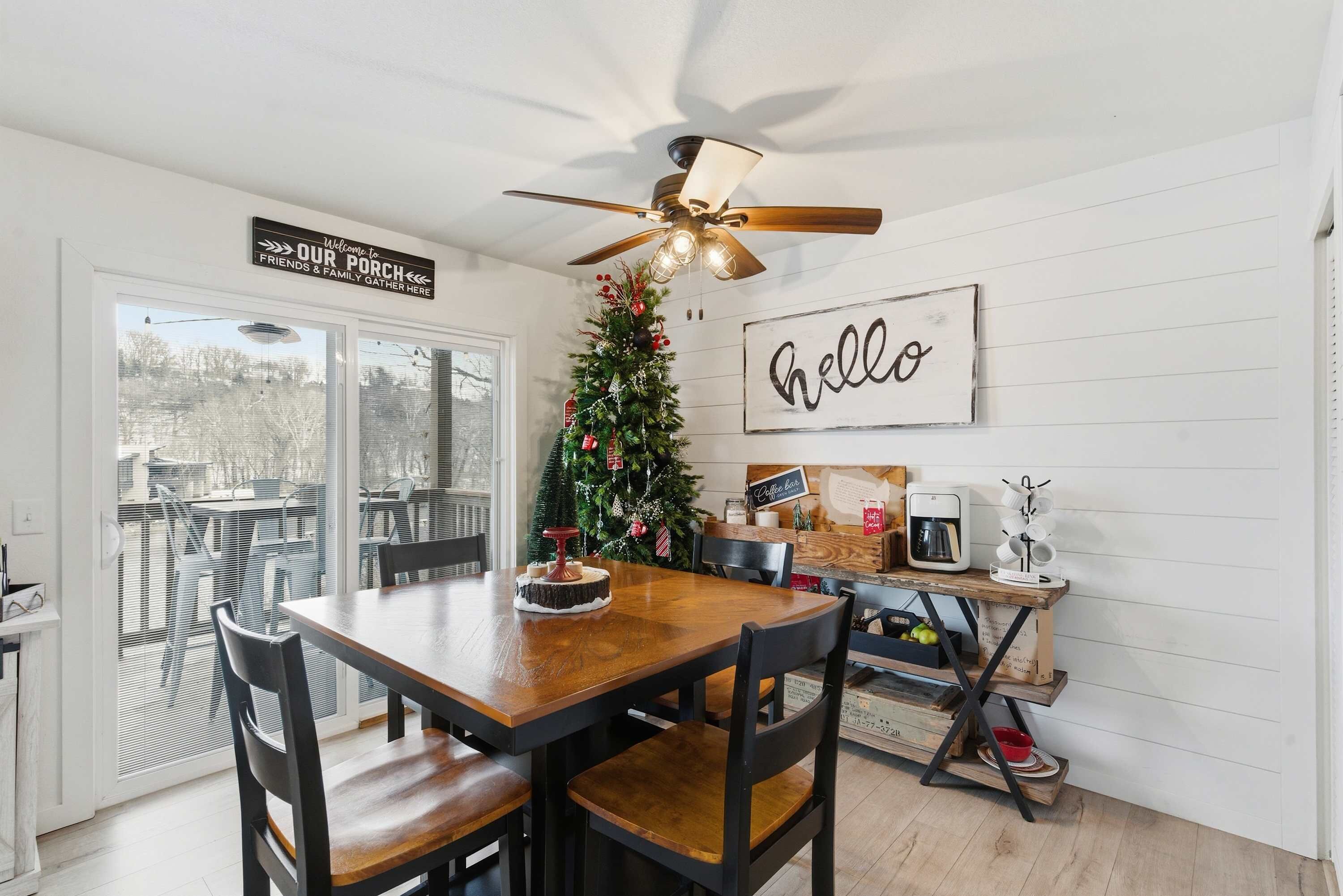 A dining area with a wooden table and chairs, a ceiling fan, a Christmas tree, and a 'hello' sign on the wall.