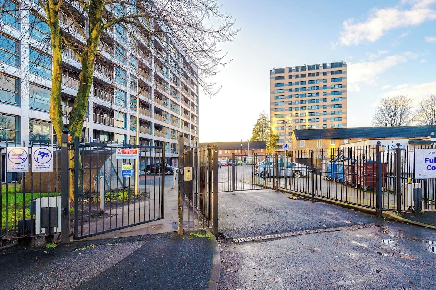 Een afgesloten parkeergebied met een gebouwtoegang, een bord waarop staat dat er cameratoezicht is, en een bord dat roken en dampen verbiedt.