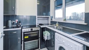 A compact kitchen with a built-in oven, a washing machine, and a window providing a view of the outside. - 1-Bed Flat | Hume | 5 Min to MMU & UoM | 15% Off (Greater Manchester)