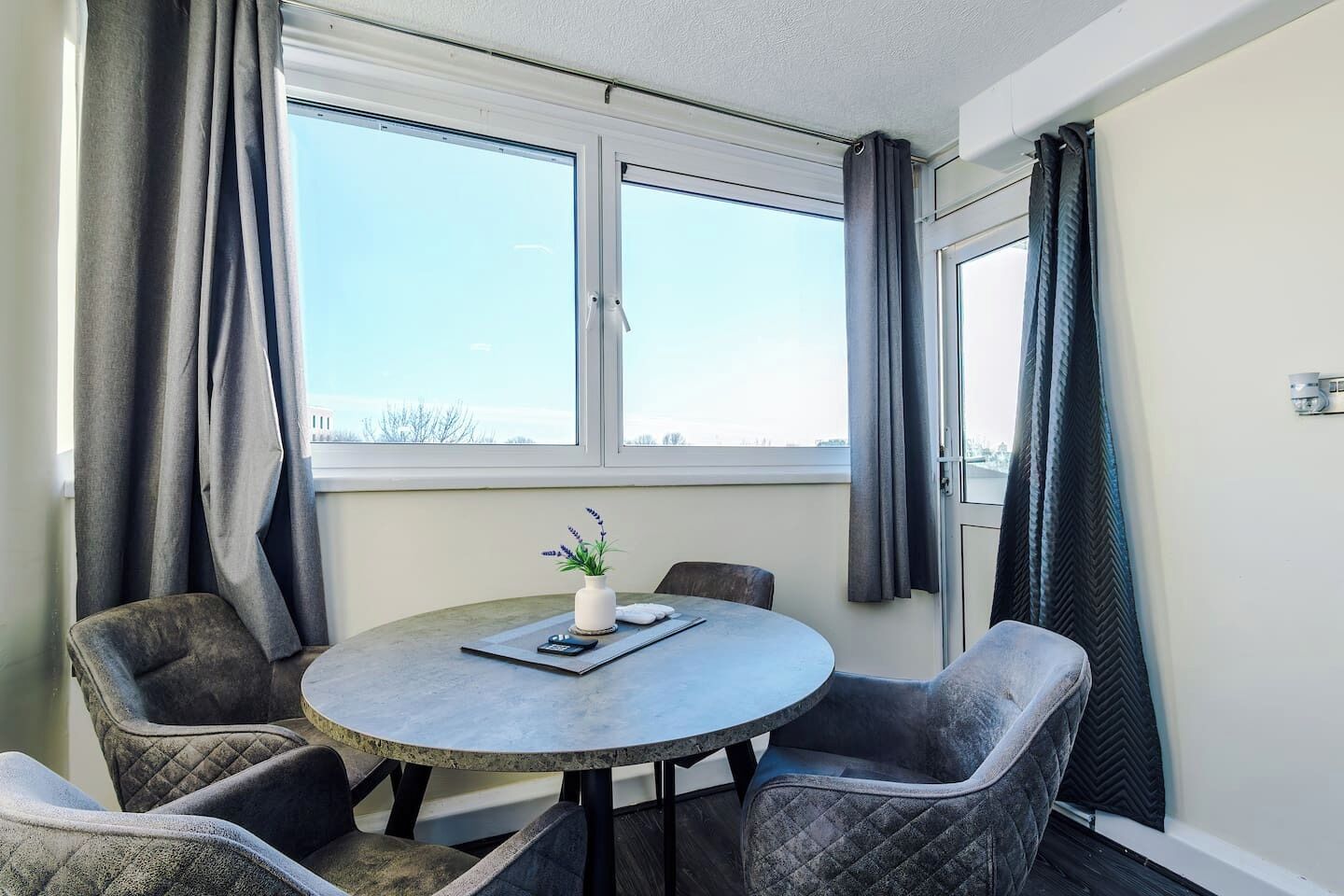 A room with a round table, four chairs, a potted plant, and a window with curtains.