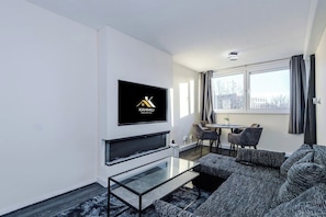 Un salon moderne avec un téléviseur à écran plat fixé au mur, une table basse en verre, un canapé gris, un coin repas avec une table et des chaises, et une fenêtre avec des rideaux.