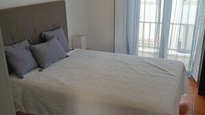 Quarto com cama, almofadas, tapete azul e vista para o exterior através de uma janela com cortinas.