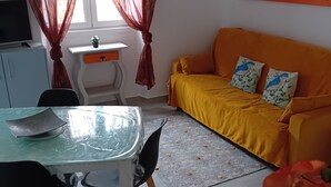 Quarto com mesa de jantar, cadeiras, sofá, televisão, escrivaninha pequena e janela com cortinas.