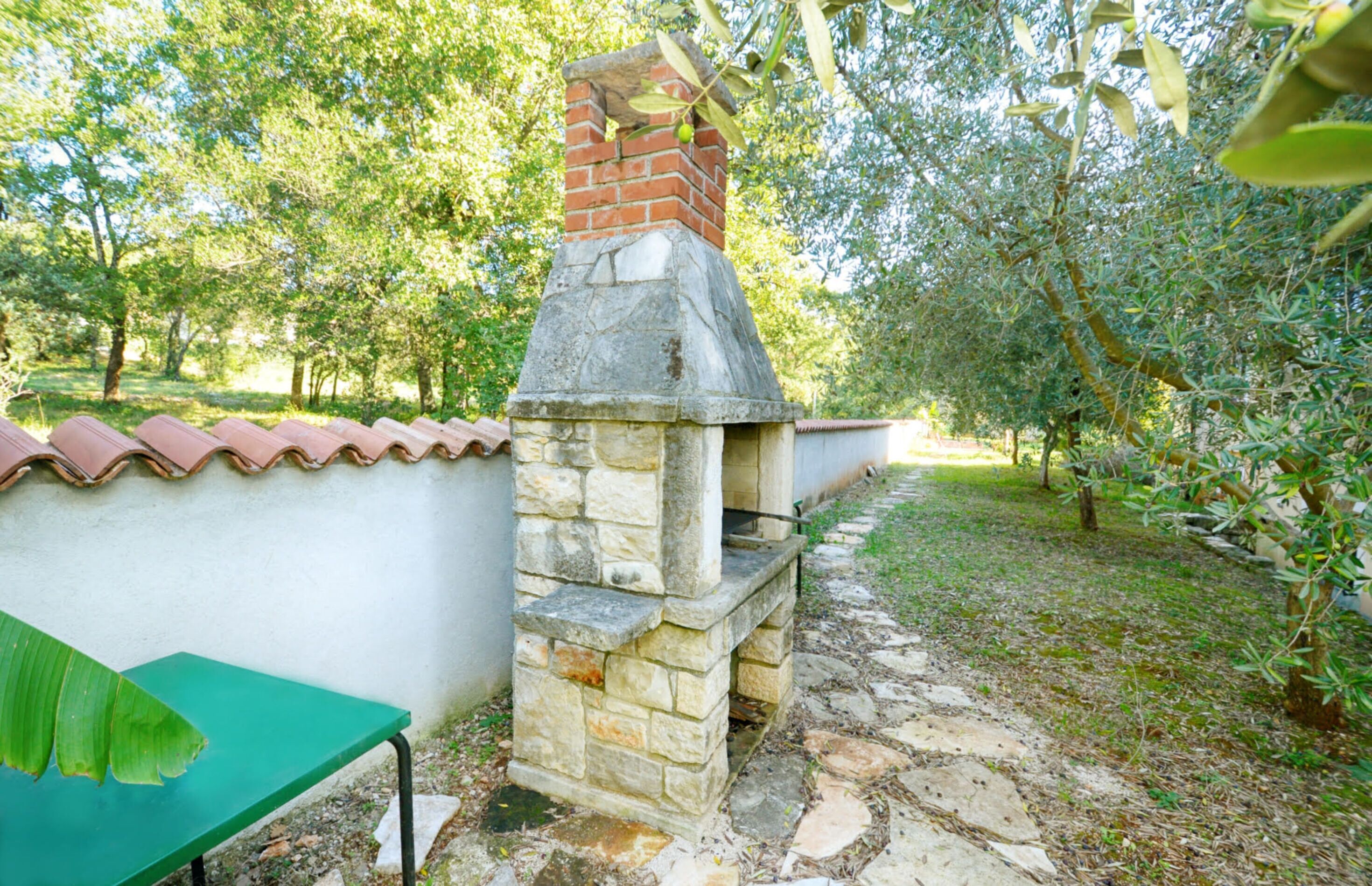 Kivestä rakennettu ulkouuni, jossa on tiilestä tehty savupiippu. Uunin ympärillä on puutarha, jossa kasvaa puita, ja sen lähellä kulkee kivipolku.