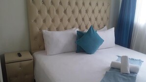 Una cama con cabecera capitonada, almohadas blancas y azules, una manta azul y un control remoto sobre un pequeño otoman junto a la cama.