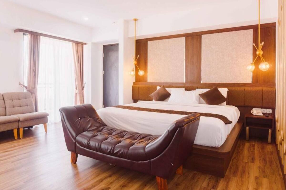 A modern hotel room with a large bed, a leather armchair, a wooden floor, and a view of the city through the window.