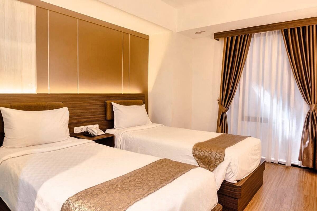 A hotel room with two beds, a wooden headboard, a bedside telephone, and a window with curtains.