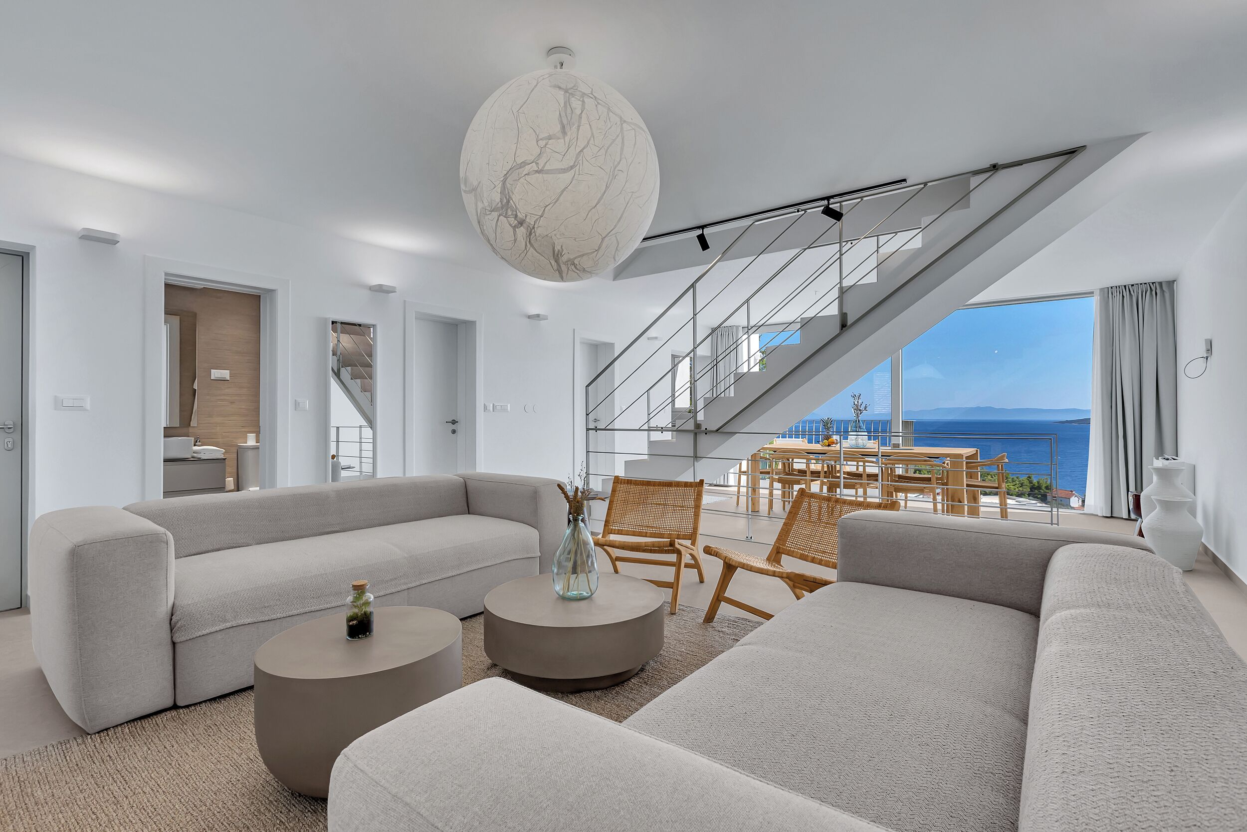 Een moderne woonkamer met een grote bank, een rond salontafeltje en rieten fauteuils. Een trap leidt naar een balkon met uitzicht op zee.