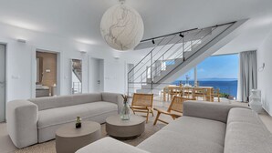 Een moderne woonkamer met een grote bank, een rond salontafeltje en rieten fauteuils. Een trap leidt naar een balkon met uitzicht op zee.
