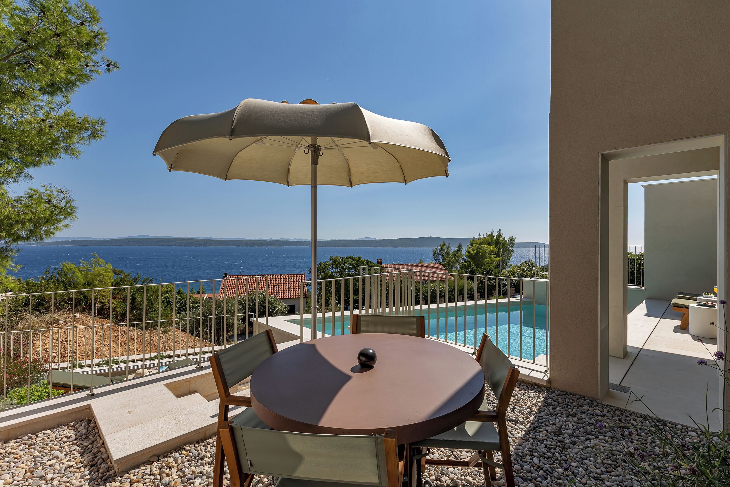 A round table with chairs and a beige umbrella, overlooking a swimming pool and the sea.