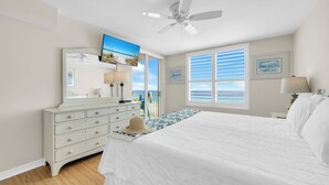 Ein Schlafzimmer mit Bett, Kommode, Deckenventilator, Fernseher und einem Fenster mit Jalousien.