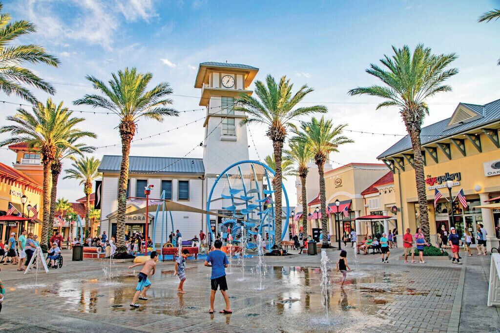 A public square with a water fountain, palm trees, and a clock tower.