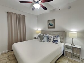 A bedroom with a bed, a ceiling fan, a nightstand, a lamp, and a wall art piece. - Tradewinds #002 | Ground-Floor, Gulf-Front, Resort Pool + Snowbird-Friendly (Orange Beach)