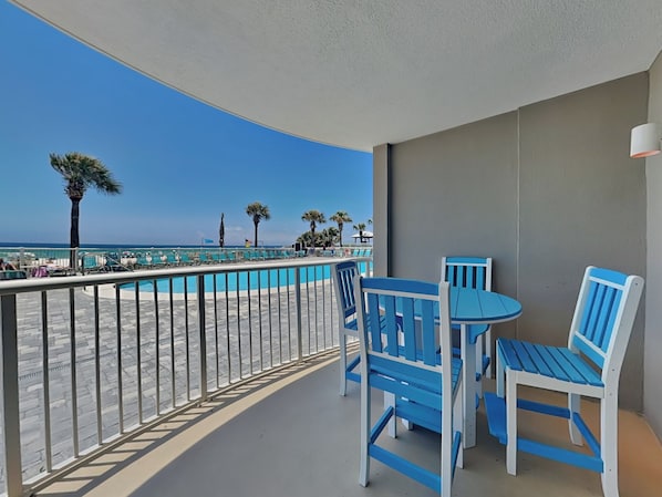 A balcony with a view of a swimming pool and beach, featuring blue chairs and a table. - Tradewinds #002 | Ground-Floor, Gulf-Front, Resort Pool + Snowbird-Friendly (Orange Beach)