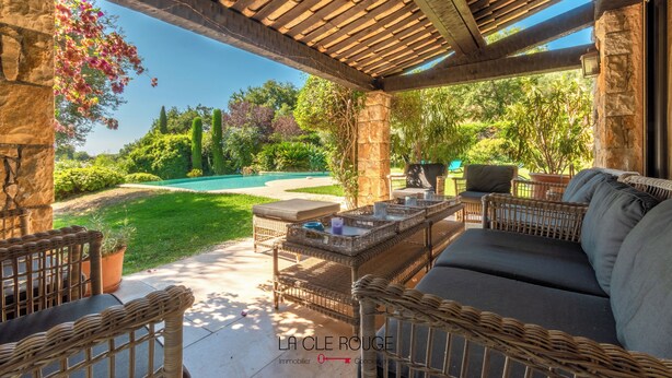 An outdoor patio with wicker furniture, a stone pillar, and a view of a swimming pool and greenery. - Villa "Les Lucioles (Vence)