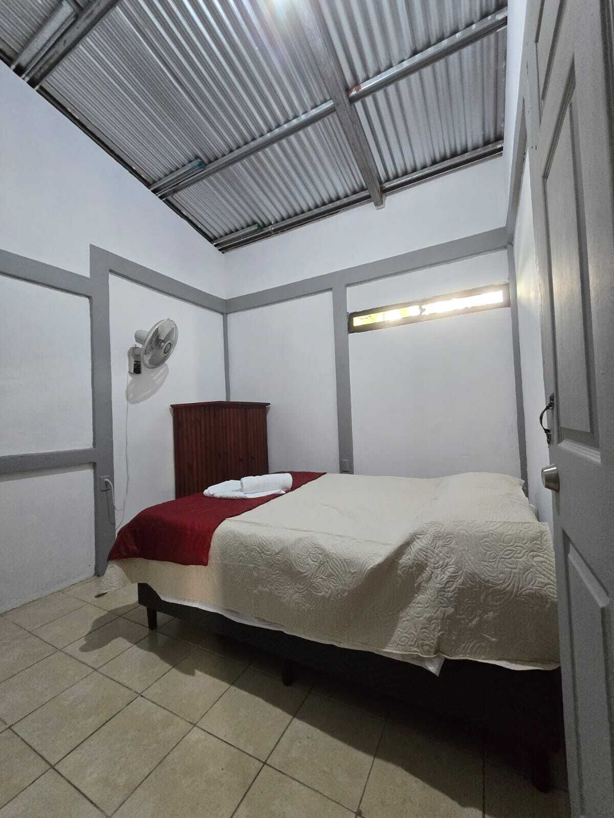 A small, single-bed room with a metal ceiling, a wooden wardrobe, a wall-mounted fan, and a bedside table.