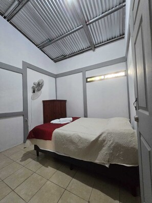 A small, single-bed room with a metal ceiling, a wooden wardrobe, a wall-mounted fan, and a bedside table. - Stay at Alexas Nature House (Arenal)