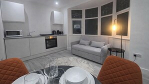 A compact kitchen with a microwave, oven, and coffee maker. There is a grey sofa and a dining table with chairs. - Luxury Studio Flat in Streatham | Close to Central London (London)