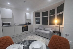 Una cocina compacta con microondas, horno y cafetera. Hay un sofá gris y una mesa de comedor con sillas.
