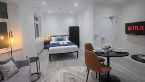 A modern hotel room with a bed, a dining table with chairs, a TV mounted on the wall, and a small side table with a lamp. - Luxury Studio Flat in Streatham | Close to Central London (London)