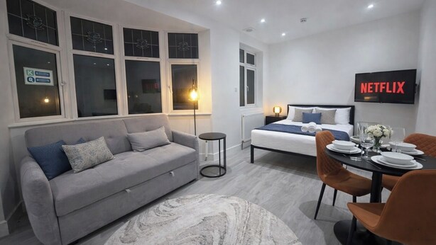 A modern bedroom with a bed, a grey sofa, a dining table with chairs, and a TV displaying 'NETFLIX'. - Luxury Studio Flat in Streatham | Close to Central London (London)