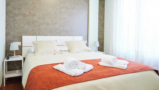 Habitación de hotel con una cama grande, ropa blanca, una manta roja y dos toallas enrolladas sobre la cama.