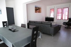 Ein modernes Wohnzimmer mit grauem Sofa, einem Esstisch mit Stühlen, einem Fernseher auf einem Schrank und einem Fenster mit roten Vorhängen.