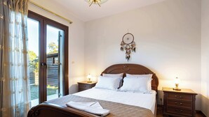 A neatly made bed with a wooden headboard, two bedside tables with lamps, a dreamcatcher on the wall, and a sliding glass door to the outside. - Villa Amorata - Schinias Private Garden Retreat (Schinias)