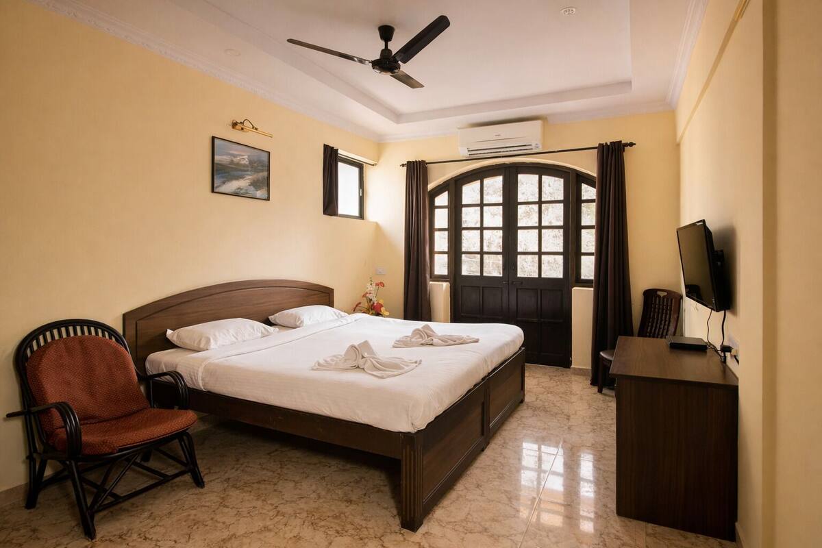 A hotel room with a bed, a chair, a desk with a computer, an air conditioning unit, and a window with curtains.
