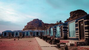 Una fila de casas móviles azul y blanco con rayas, con ventanas grandes, frente a un paisaje desértico y una destacada formación rocosa al fondo.