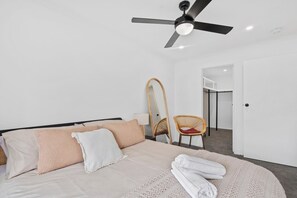 A bedroom with a bed, a ceiling fan, a chair, a mirror, and a door leading to another room. - Palm-Feel Townhouse in the Heart of Burleigh (Burleigh Heads)
